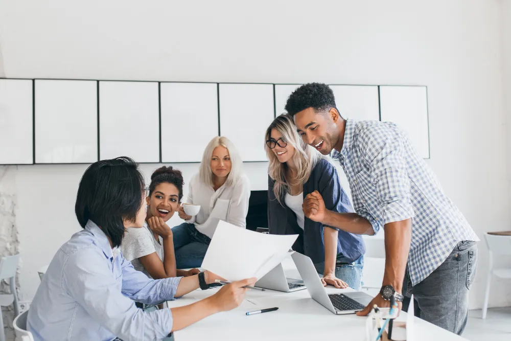 Diverse team gathers around a white desk, enthusiastically reviewing papers and laptops together.