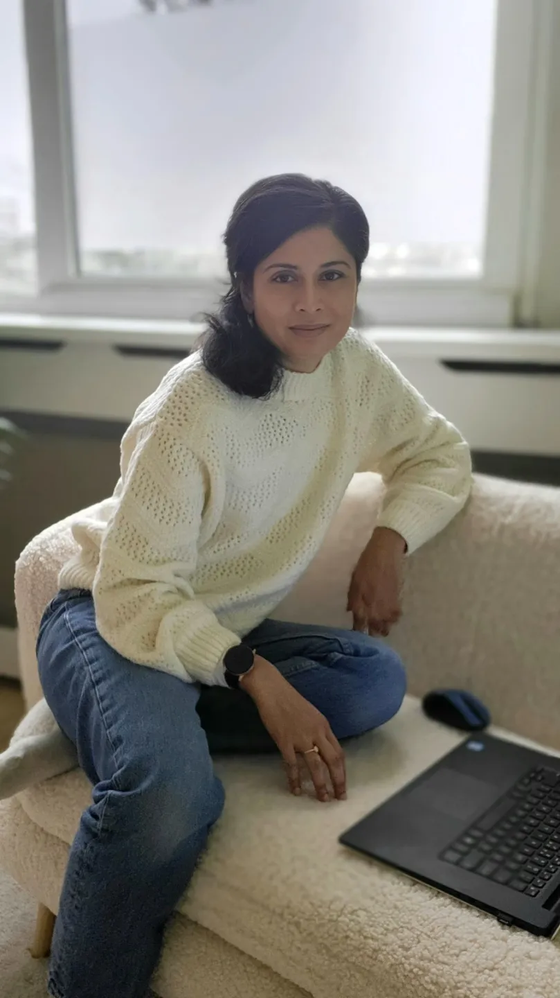 Woman in a cream sweater perched on a couch arm, looking at the camera beside an open laptop.