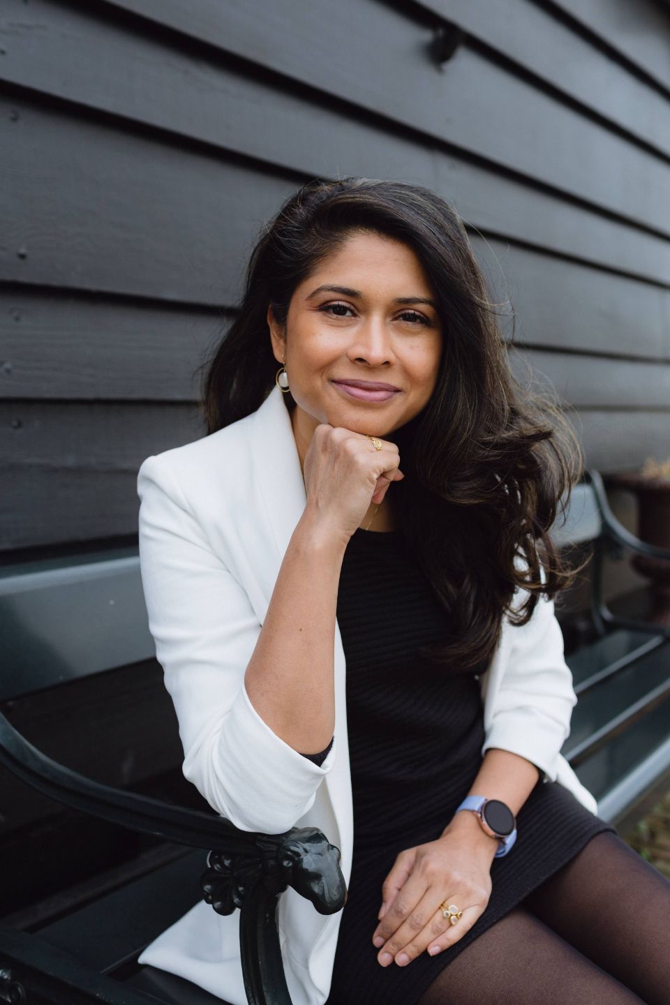 Professional woman in a white blazer sits on a bench, chin resting on her hand in a relaxed pose.